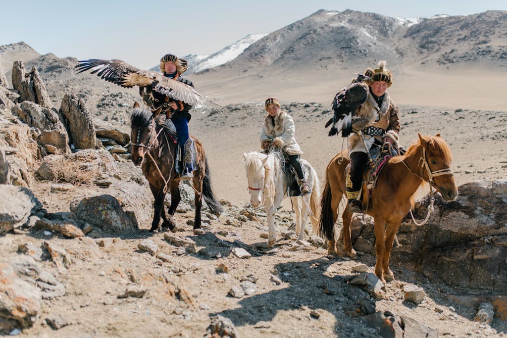 Full body Mongolian horsemen wearing traditional local clothes carrying eagles on hands and riding horses along spacious rocky terrain during eagle hunting